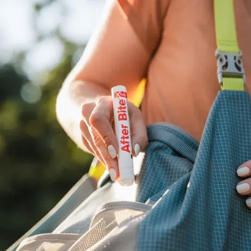 Person with white painted nails holding a white and red marker labeled "After Bite" outdoors, with a yellow strap of a backpack and a blue checkered fabric visible.